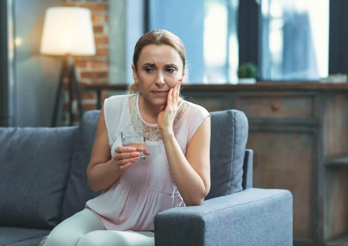 Woman holding a glass of water and touching her cheek while experiencing tooth discomfort related to needing a root canal and crown.